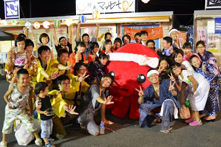 南三陸の夏祭り会場で、大きな赤いタコのキャラクター（オクトパス君）を囲んで浴衣姿の子どもや大人たちが笑顔で写っている集合写真