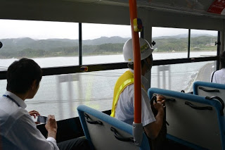 バスの車内で作業服を着た人と他の乗客が窓の外に広がる海の景色を見ている様子の写真