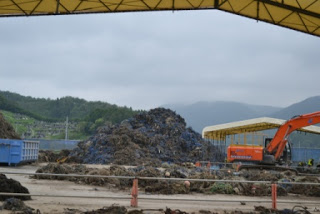 曇り空の下に緑の丘や山が広がり、右奥に白い屋根のテント、左側に青いコンテナが2つあり、大量の瓦礫が積まれ、右側のオレンジ色のショベルカーで作業を行っている様子の写真