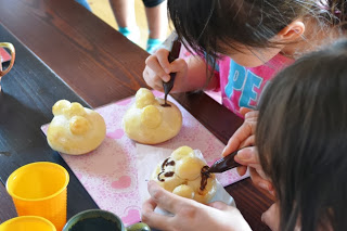 女の子たちがテーブルで丸い3つの粒が乗った丸いパンの表面に、チョコペンで顔を描いている写真