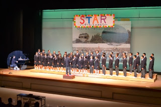 制服を着た中学生の合唱団がステージ上に整列し、指揮者の指導のもと合唱を披露しており、左側ではピアノ伴奏をしている生徒の姿も見える写真