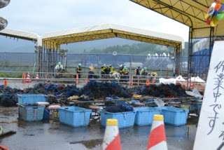 交通コーンやフェンスが配置された間に、瓦礫が入れられた青い容器が置かれ、奥の白い大きなテントの下で、ヘルメットと安全ベストを着用した作業員が複数人おり、魚網の処理を行っている様子の様子