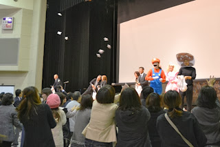 ステージ上でユニークな仮装をした数人の大人たちが、観客に向かって小さな包みを楽しそうに投げており、観客席の人々が手を伸ばして受け取ろうとしている様子を写した写真