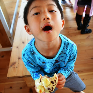 青い長袖シャツの男の子が、かじったパンを手に持ち、周りにチョコレートがついた口を大きく開けてカメラに見せている写真