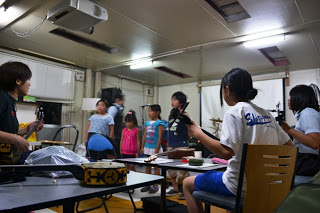 子どもたちが室内で三線の練習をしていて、前方の椅子に座った子どもが三線を弾き、後ろで他の子どもたちが立って歌を歌っている練習風景の写真