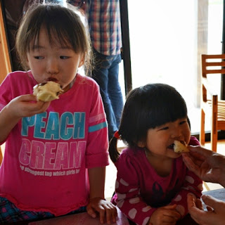 ピンクの服の女の子がパンを食べている隣で、濃いピンクの服の小さな女の子が嬉しそうにパンをかじっている写真