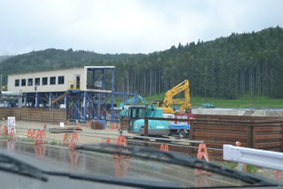 奥には木々と丘が広がり、道路沿いにはバリケードが設置され、左側に足場と支柱が組まれた解体中の窓が沢山ある駅舎のような建物があり、中央には黄色と青色のショベルカーが作業をしている工事現場の写真