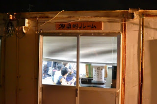 「多目的ルーム」と書かれた看板が掲げられている建物の外観で、夜間に灯りがともり、窓から子どもたちが三線の練習をしている様子が見えている写真