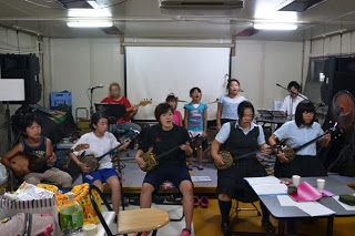 室内のステージ前で三線を持った子どもたちが座り、後ろで立って歌う子どもたちがいる、三線教室のグループ練習中の写真