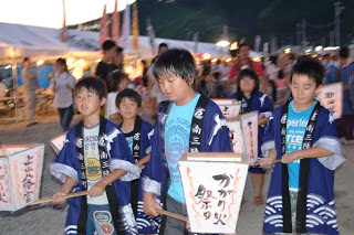 夜の祭り会場で、数人の子どもが伝統的な祭り衣装を着て提灯を持ち、祭りの行列やイベントに参加している様子が写っている写真
