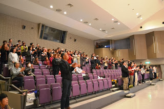 ホールの観客席で多くの観客が立ち上がり、拍手をしながら舞台上の発表に応えている様子を写した写真