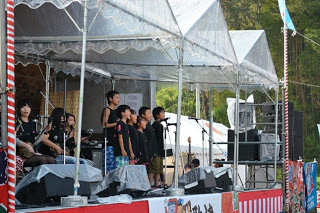 屋外ステージで黒い服を着た子どもたちの合唱グループがマイクの前に並び、その隣で三味線を演奏する子どもたちもいて、観客に向かって歌と演奏を披露している様子の写真