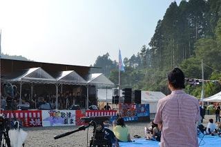 観客席から屋外ステージを撮影した写真で、ステージ上の子どもたちの合唱グループと、その前で観客が鑑賞している全景の写真