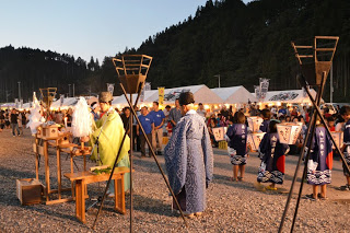 夜の祭りで、神職が神事を行う祭壇の前に立ち、祭り衣装を着た子どもたちや参加者が周囲に集まっている、地域の伝統行事の様子が写っている写真