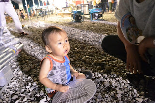 小さな子どもが手に扇子を持ち、夜の砂利の上に座って夏祭りの様子を見つめている写真