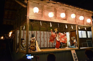 神社の舞台で、華やかな衣装をまといお面をかぶった舞手が鈴や扇を手に持ち、提灯の灯りに照らされながら神楽を舞っている様子の写真