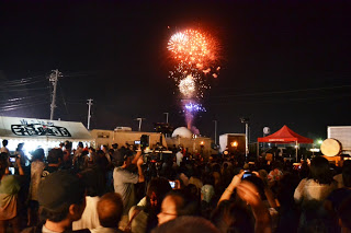 夜空に打ち上げられた色とりどりの花火を多くの観客がスマホやカメラで撮影しながら見上げている様子の写真