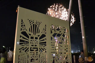 花火が打ち上げられる夜空を背景に、和風の彫刻が施されたきりこの看板の前で人々が花火を眺めている様子の写真