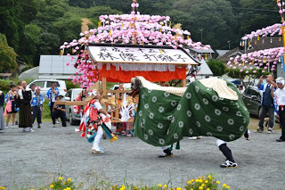 昼間の神社の境内で、色鮮やかな花飾りがついた神輿の前で獅子舞が披露されている様子の写真