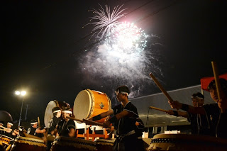 和太鼓を叩く複数の演奏者が夜空に打ち上げられた花火を背にして力強く演奏している様子の写真