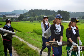 女性たちが長靴を履いて農道を歩きながら、農作業に使う木材や道具を手に運んでいる農業体験中の写真