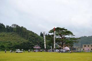 広い田んぼの中の道を、白装束の人々がのぼり旗や神輿を担ぎながらねり歩き、見物人や参加者が周囲に集まっている様子を写した写真