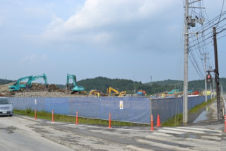 背景に丘と曇り空が広がる、車が走行している道路が交差する角地に、青と白のフェンスに囲まれた工事現場で複数の重機が作業している写真