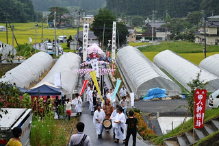 白装束を着た人々が太鼓を鳴らしながら農業用ビニールハウスが両側にある、神社までの道を練り歩いている祭りの様子の写真