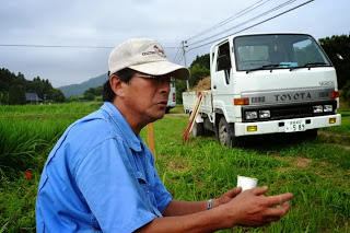 田んぼの脇に座っている青い作業着と白い帽子をかぶった阿部博之さんの写真