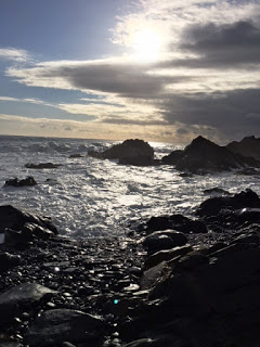 波が岩に打ち寄せる海岸の水面を、雲間から差す光が照らしている様子の写真