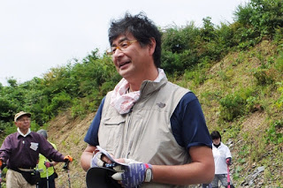 ベージュ色のベストを着た男性が草木が生えた斜面の前でメモを手に話をしている写真