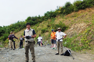 数人の男性が右側に土と草の斜面がある山道の広場で休憩をしている様子を写した写真