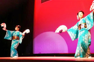 白い柄の入った緑色の着物を着た二人の女性が、赤い背景のステージで扇子を使って舞を披露している写真