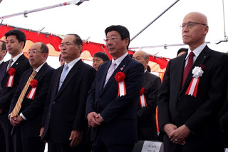 室内の式典会場で、赤白の幕の前に立っている複数の男性がスーツを着て胸に赤や白のリボンを付けて整列しており、厳粛な雰囲気の中で式典に参加している写真