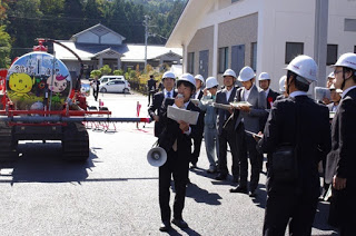 屋外でスーツにヘルメットをかぶった多くの人たちが、色鮮やかなデザインのタンクを載せた車両の前で説明を聞いており、女性がメガホンを使って案内している写真