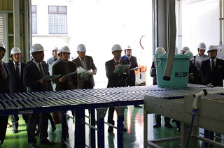 屋内の工場でヘルメットをかぶった人々がコンベアと青い容器を見学しており、資料を手に説明を聞いている写真