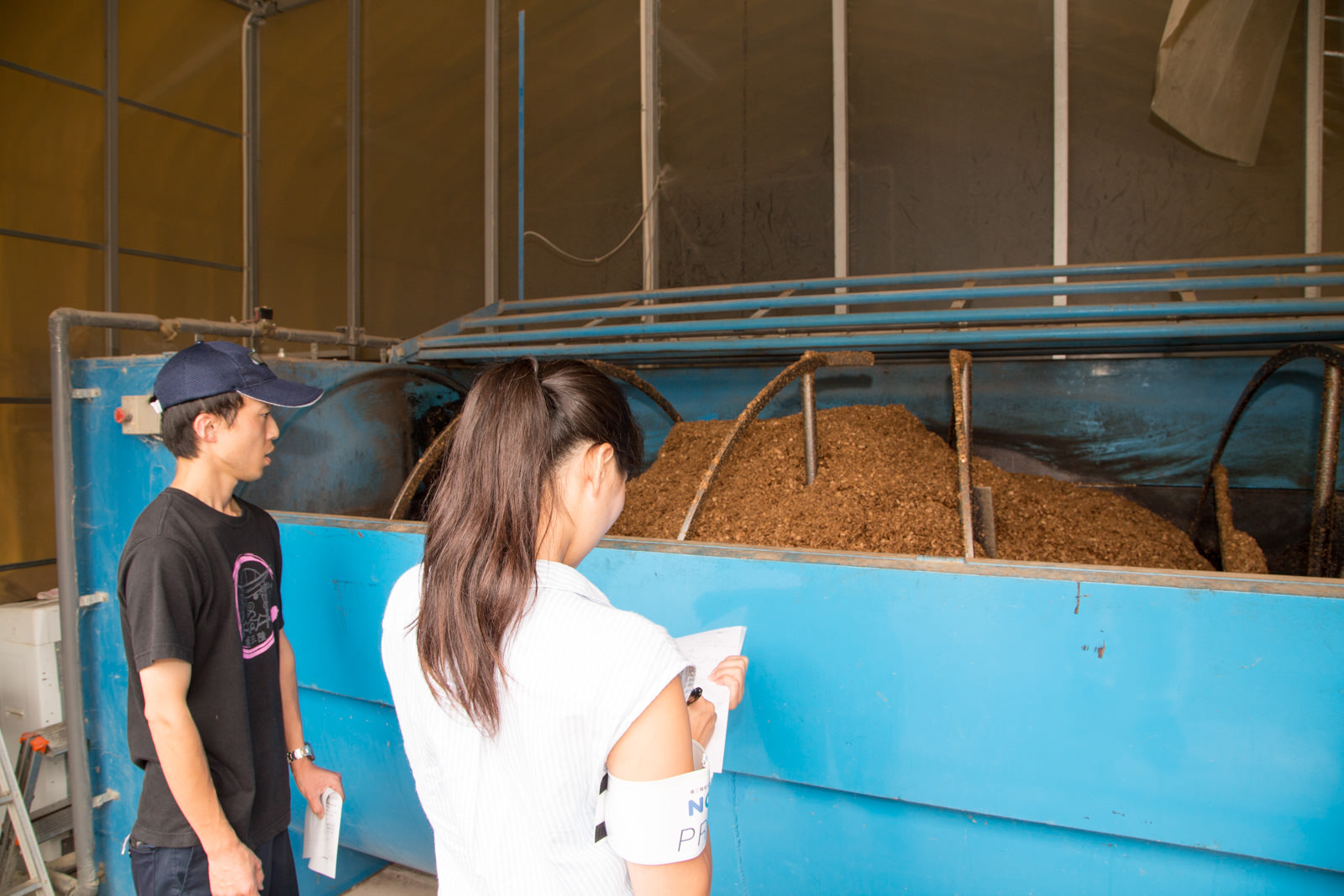 大きな青い機械の前で、男性スタッフがおが粉を見ながら説明しており、女性が聞きながらメモをとっている写真