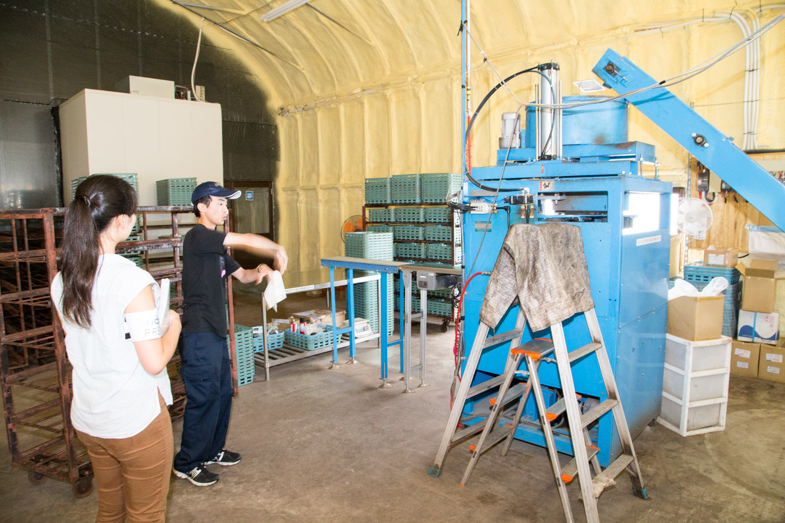 青色の機械が置かれた作業場で、キャップを被った男性が女性に向かって説明をしている写真