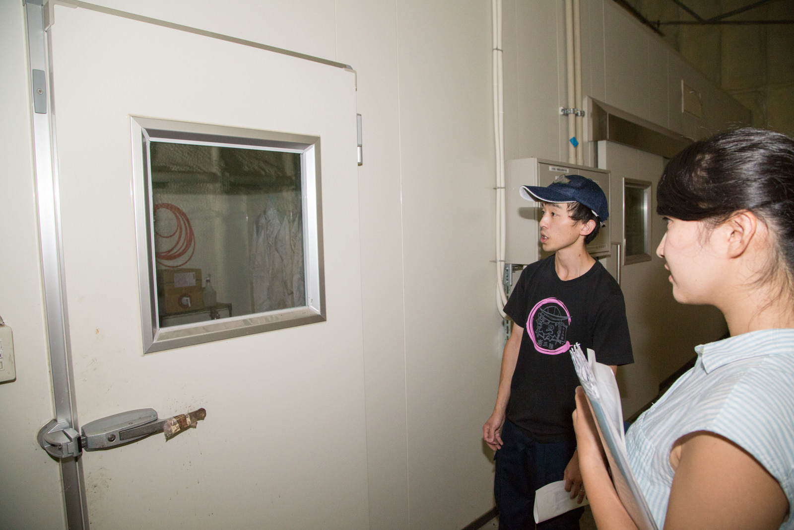 白い壁の密閉された部屋の前で、男性が女性に向かって施設内部について説明している様子の写真