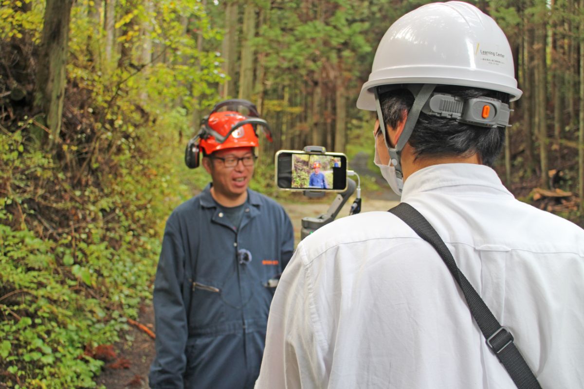 森林の道で白いヘルメットとマスクを着用した男性が、赤いヘルメットを被り作業服姿の男性に向けてスマートフォンで撮影をしている様子の写真