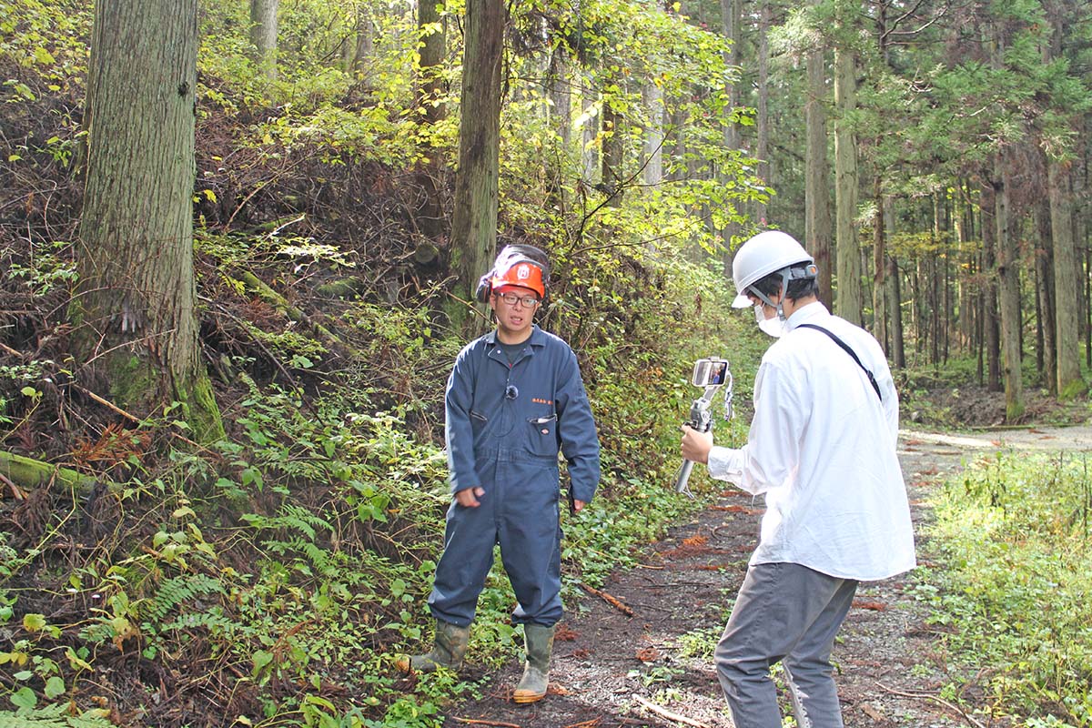 森の中でヘルメットをかぶり作業服を着た男性がカメラを持った人物に撮影されている様子の写真