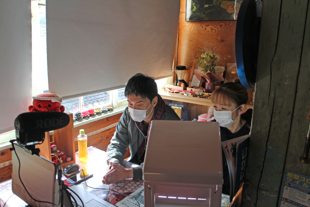 外部マイクが設置されたテーブルに並んで座ったマスク姿の男性と女性がパソコンを操作している写真