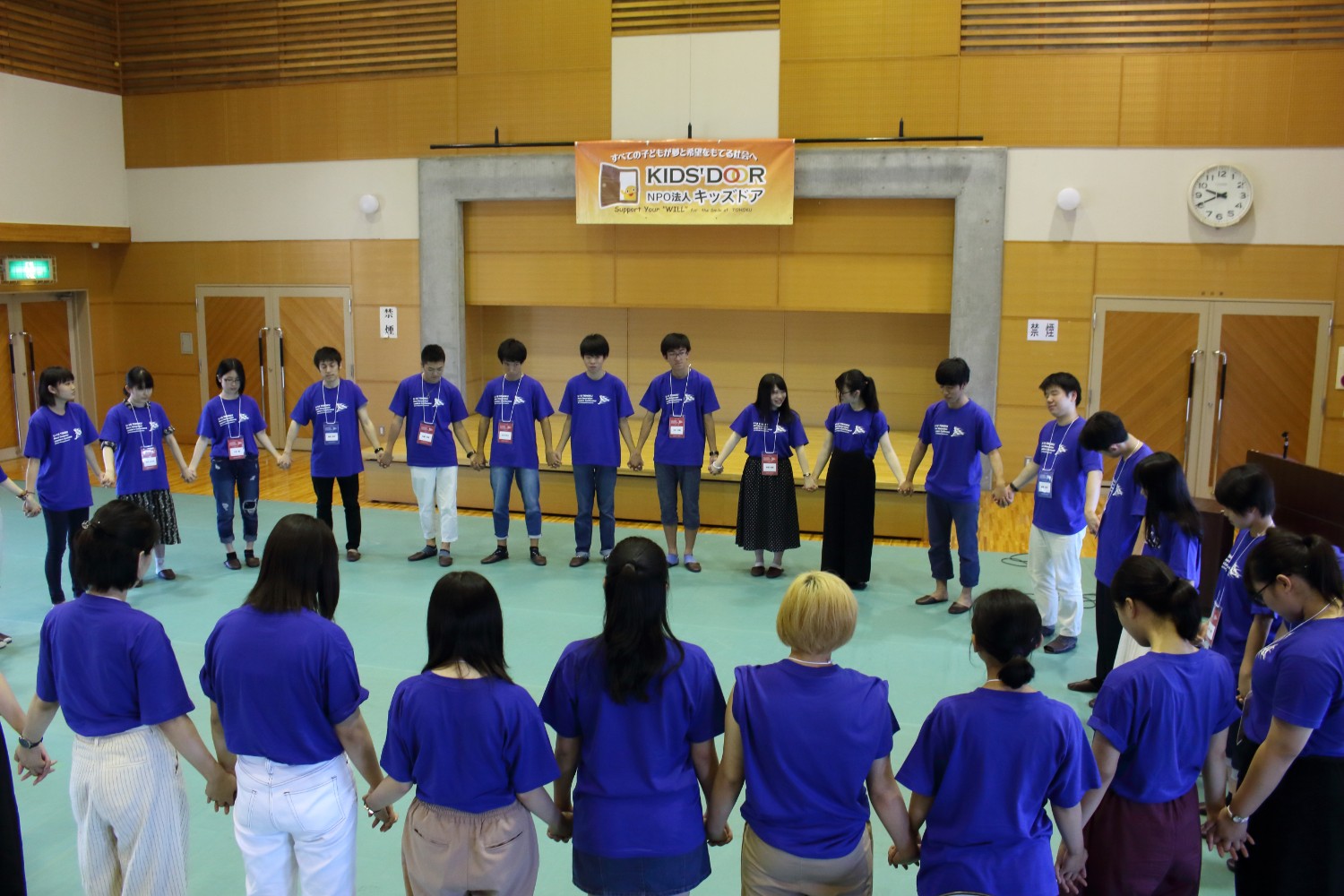 「KIDS' DOOR」という横断幕が掲げられた体育館のような講堂で、青いお揃いのTシャツを着た十数人の若者たちが手をつなぎ一つの輪を作って、心を合わせている様子の写真