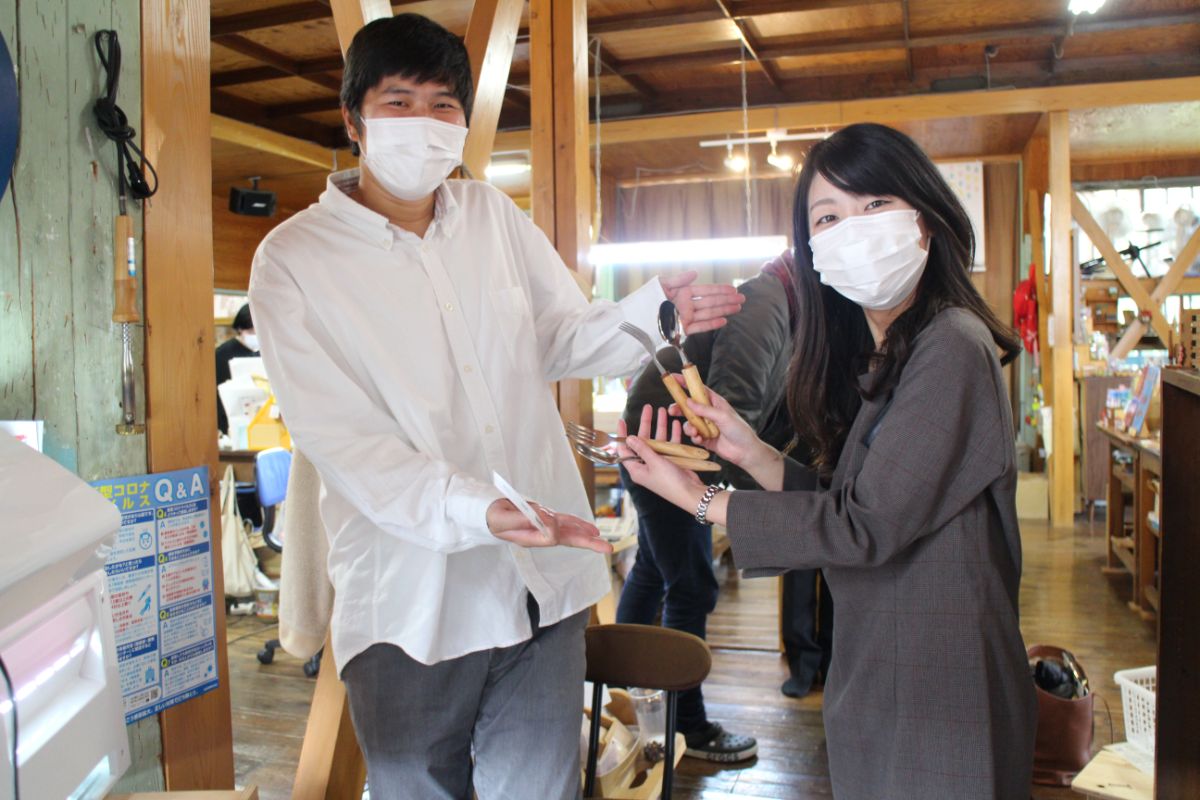 室内でマスクをした男女二人が木製のフォークとスプーンを手に持ち、笑顔で紹介している様子の写真