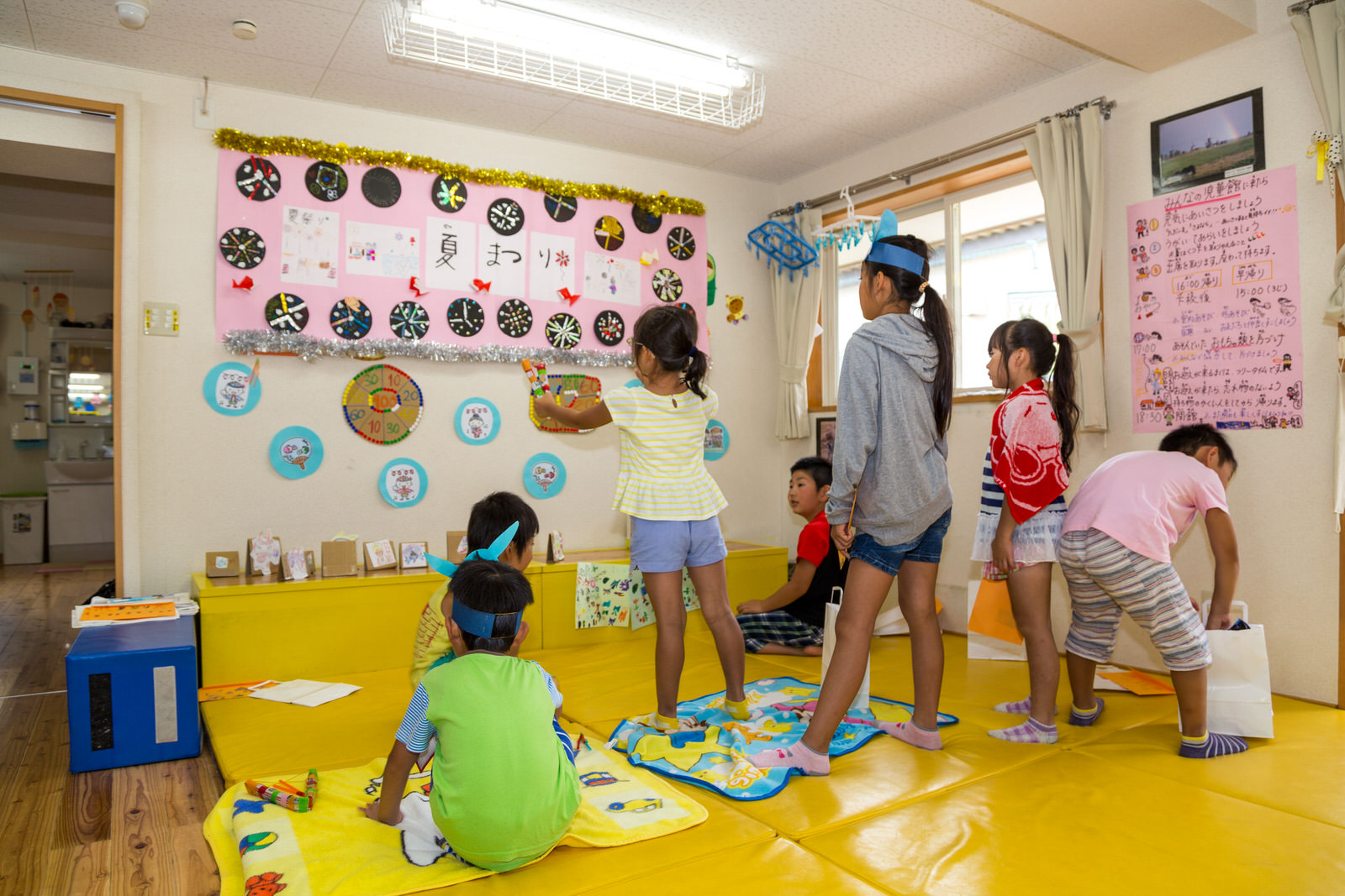 1人の女の子が「夏まつり」と書かれた壁飾りの下に貼った的をめがけて的当てをしており、その周りで複数の子どもたちが順番待ちをしている様子の写真