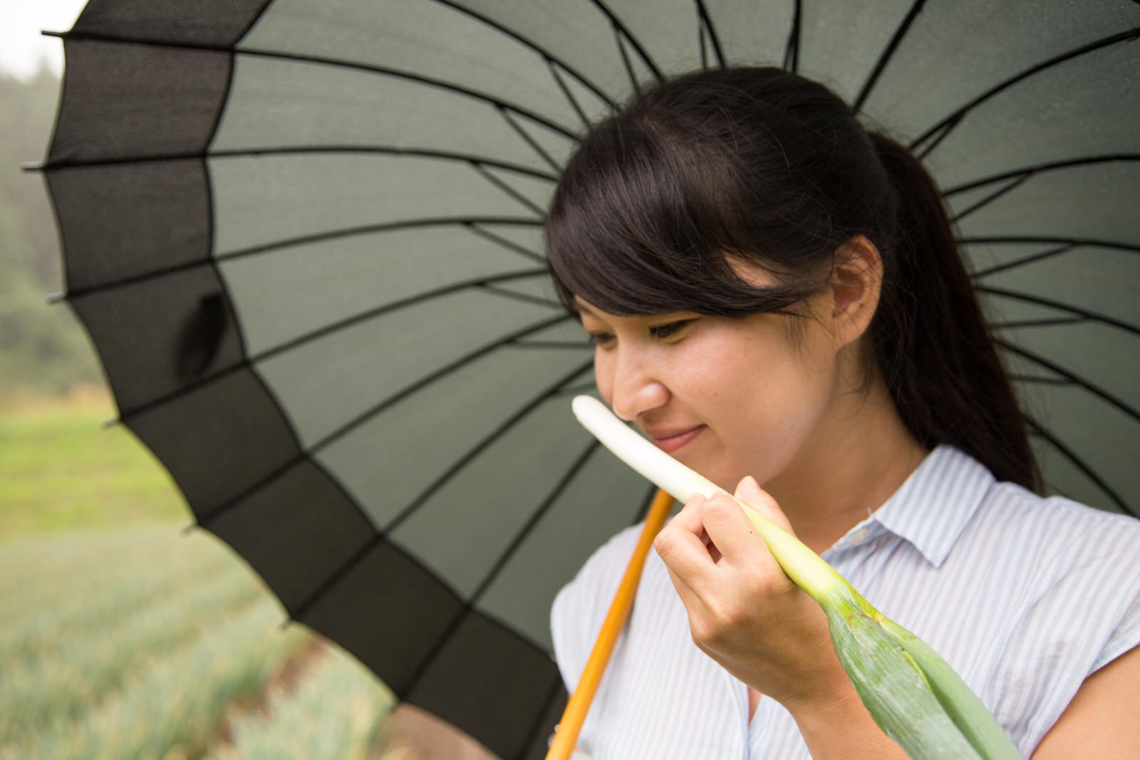 大きな黒い傘をさした女性が、手に持った長ネギの香りを嗅いでいる様子の写真