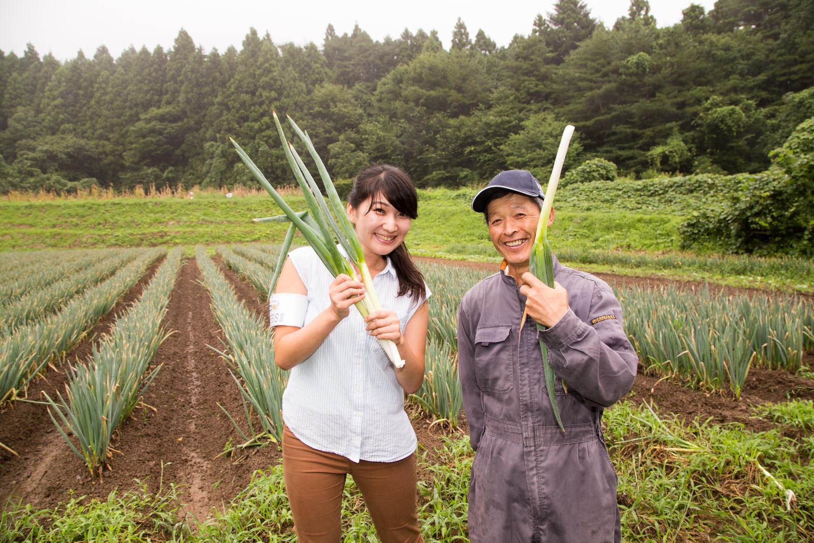 畑の中で女性と笑顔の作業着姿の男性が並び、手に収穫した長ネギを持ちながらポーズを取っている写真