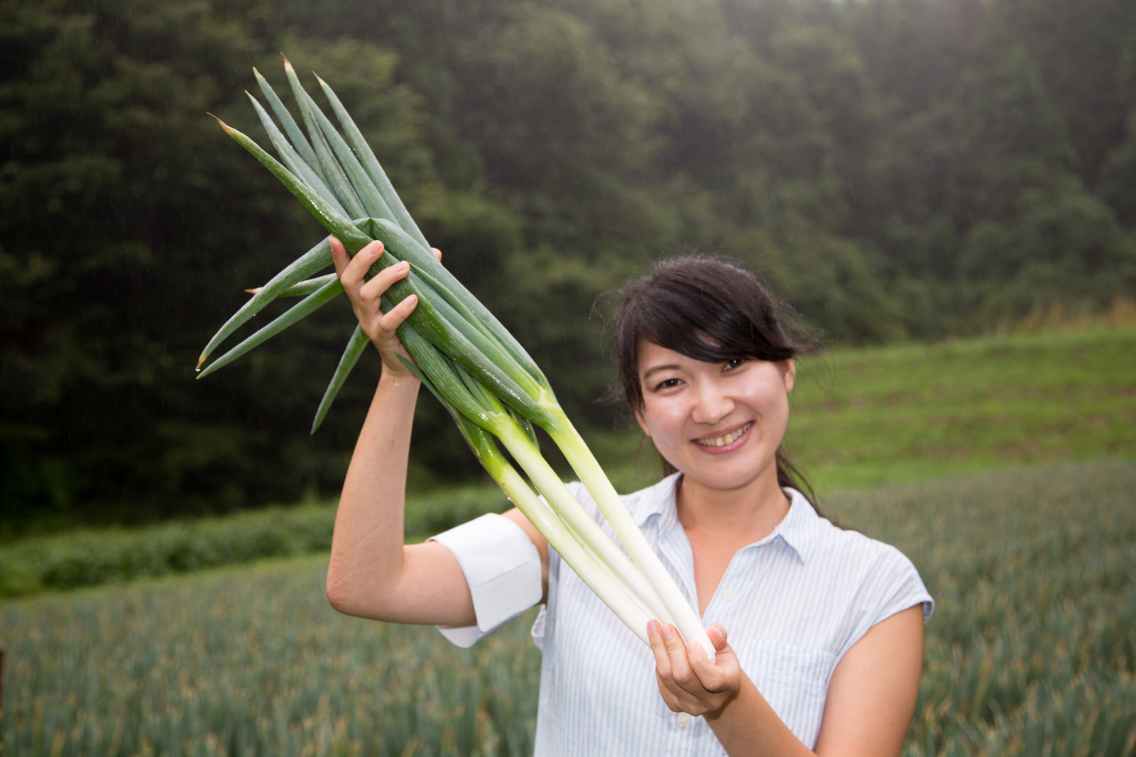 女性が大きく育った長ネギの束を両手で高く持ち上げて笑顔を見せている様子の写真