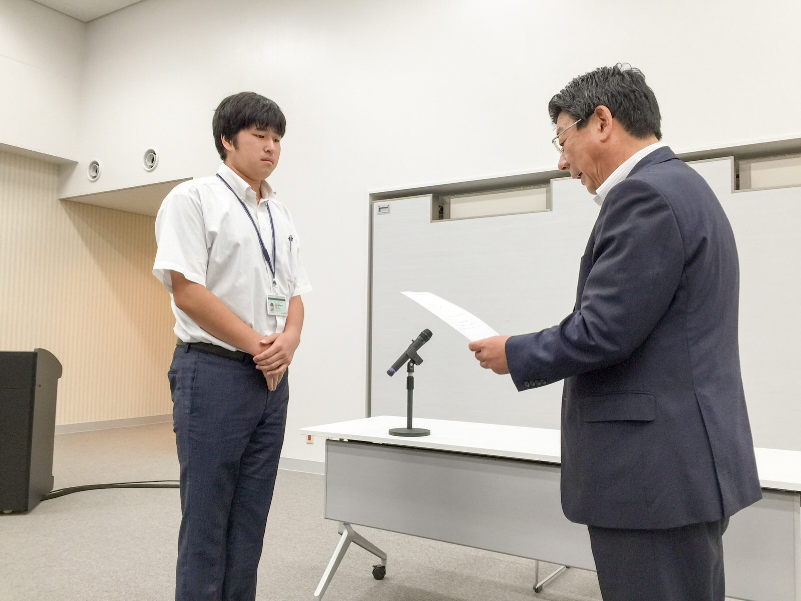 マイクが置かれた白い机の前で、白いシャツを着た学生と向かい合って立つスーツ姿の佐藤町長が、指示書を読んでいる様子の写真