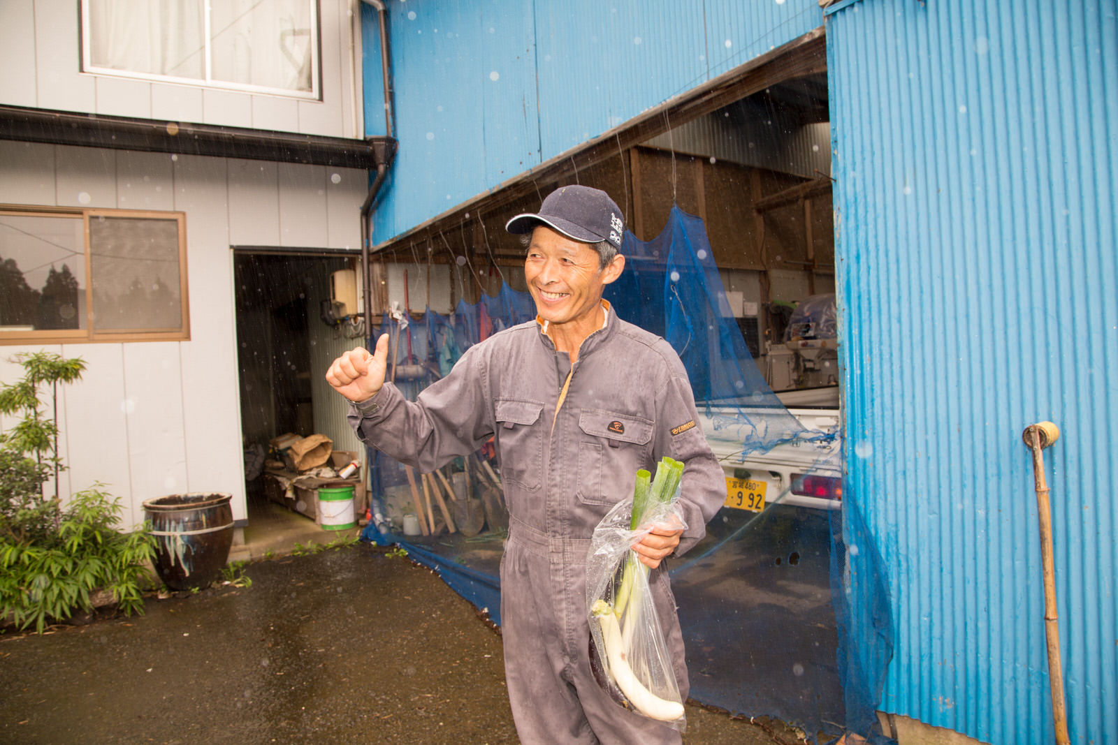 作業着を着た男性が家の前で長ネギの入った袋を手にし、笑顔で親指を立てている様子の写真